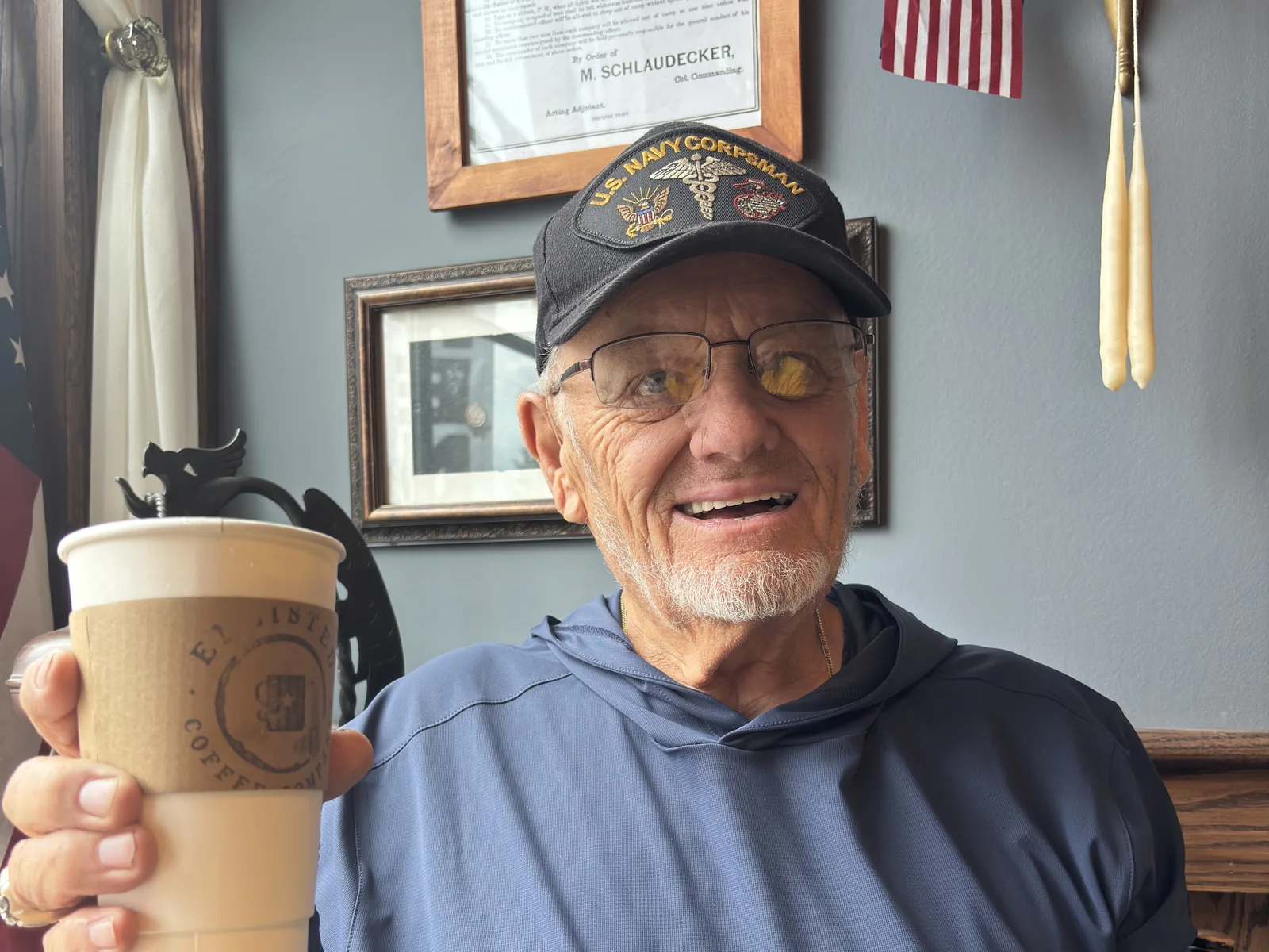 Dennis Myers smiles and holds up a paper coffee cup while seated inside Enlisted Coffee in Waterford, Pennsylvania.